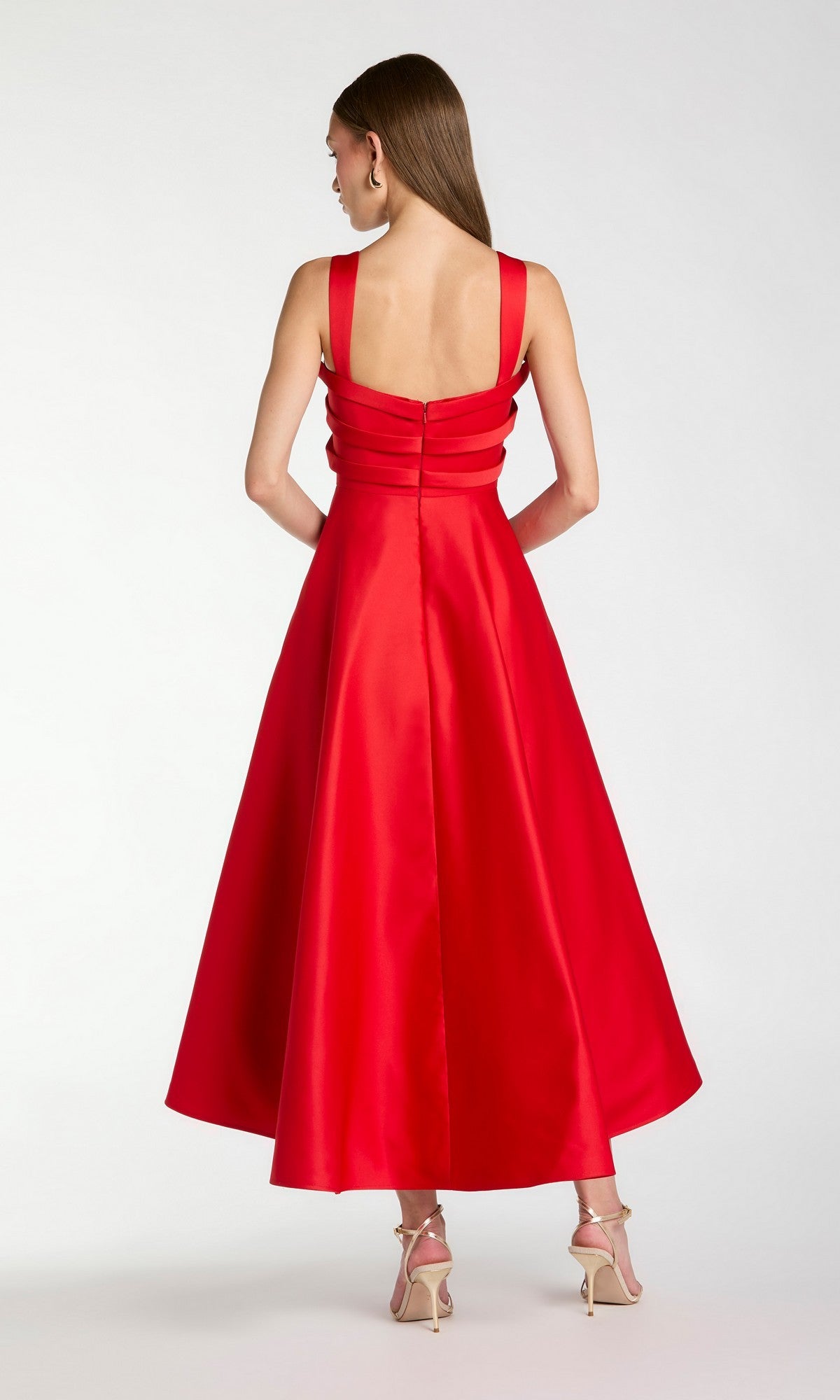 A woman stands facing away, wearing the Mac Duggal 21247 tea-length party dress—an elegant sleeveless, backless red satin cocktail dress with wide straps—paired with beige high heels against a plain light background.